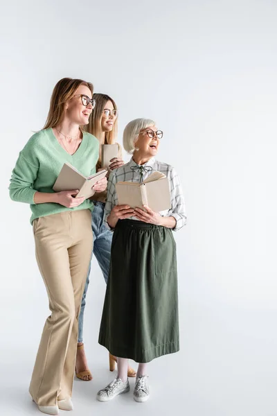 В полный рост три поколения улыбающихся женщин в очках с книгами на белом — стоковое фото