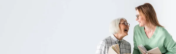 Madre mayor riendo con hija en gafas mientras sostiene libros aislados en blanco, pancarta - foto de stock