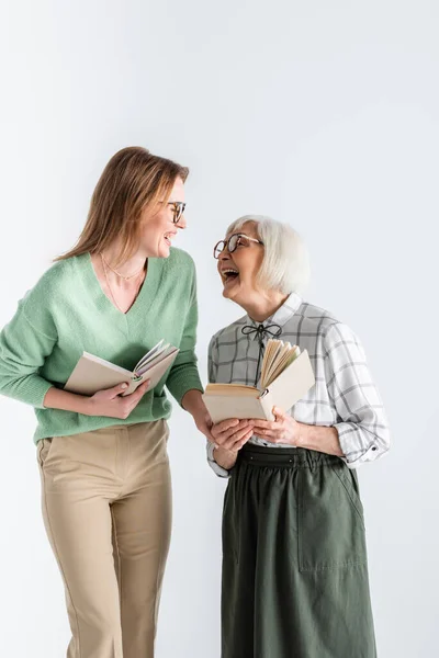 Mãe sênior rindo com a filha em óculos enquanto segurando livros isolados em branco — Fotografia de Stock