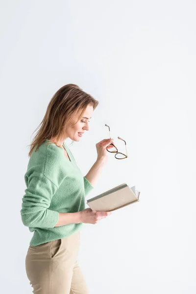 Mujer alegre sosteniendo gafas y leyendo libro aislado en blanco - foto de stock
