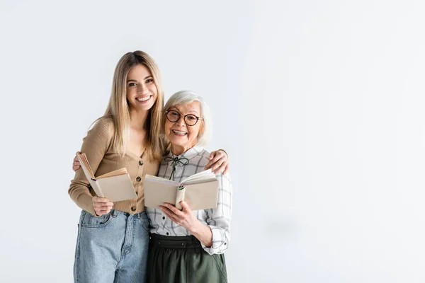 Happy young woman hugging granny in glasses and holding books isolated on white — Stock Photo