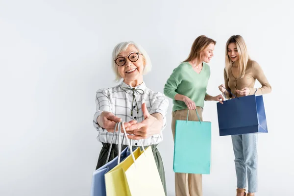 Cheerful senior woman holding shopping bags near daughter and granddaughter on blurred background — Stock Photo