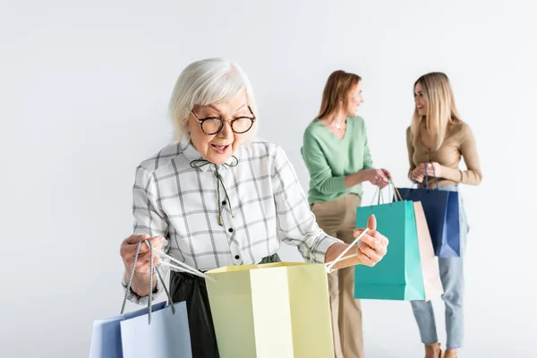 Surprised senior woman looking at shopping bag near daughter and granddaughter on blurred background — Stock Photo