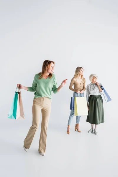 Полная длина счастливой женщины, держащей сумки с покупками и указывая пальцем рядом с дочерью и старшей матерью на размытом белом фоне — стоковое фото