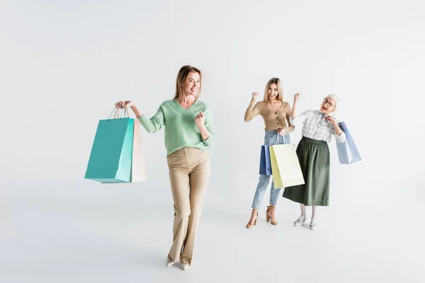 Longitud completa de la mujer feliz con bolsas de compras cerca de hija y madre mayor sobre fondo blanco borroso - foto de stock