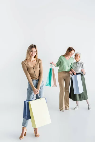 Comprimento total de feliz jovem mulher de pé com sacos de compras perto de mãe e avó no fundo borrado — Fotografia de Stock