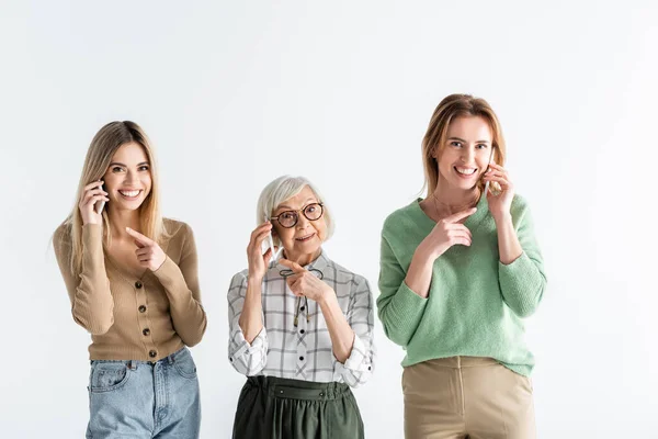 Tres generaciones de mujeres felices hablando y señalando con los dedos a los teléfonos inteligentes aislados en blanco - foto de stock