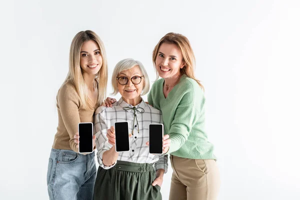 Tres generaciones de mujeres alegres sosteniendo teléfonos inteligentes con pantalla en blanco aislado en blanco - foto de stock