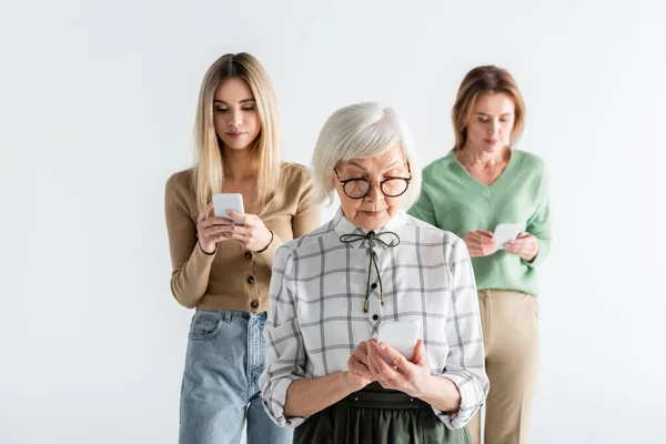 Senior woman in glasses using smartphone near daughter and granddaughter on blurred background — Stock Photo