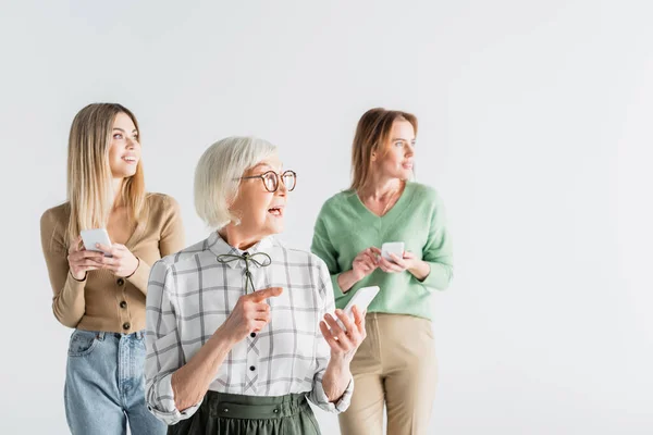 Verblüffte Seniorin mit Brille, Smartphone in der Hand und Finger in der Nähe von Tochter und Enkelin auf verschwommenem Hintergrund — Stockfoto