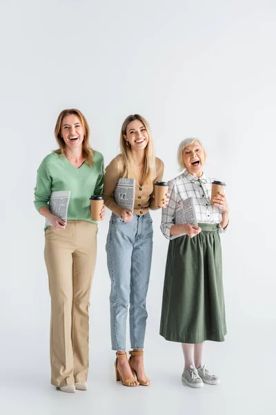 Longitud completa de tres generaciones de mujeres alegres sosteniendo periódicos y vasos de papel en blanco - foto de stock