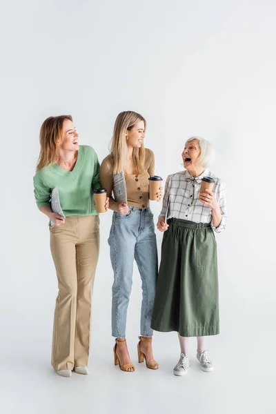 Full length of three generation of smiling women holding newspapers and paper cups on white — Stock Photo