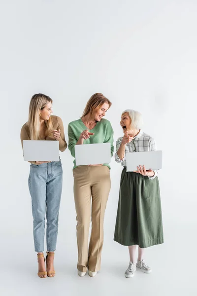 Longitud completa de tres generaciones de mujeres alegres señalando con los dedos, mirándose unos a otros y sosteniendo computadoras portátiles en blanco - foto de stock