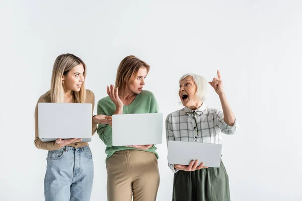 Madre e hija joven sosteniendo computadoras portátiles y mirando a la mujer mayor señalando con el dedo aislado en blanco - foto de stock