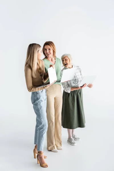 Longitud completa de tres generaciones de mujeres sosteniendo computadoras portátiles y sonriendo en blanco - foto de stock