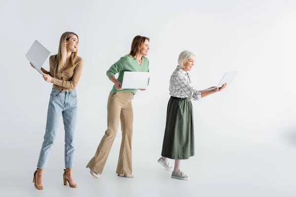 Pleine longueur de trois générations de femmes joyeuses tenant des ordinateurs portables sur blanc — Photo de stock