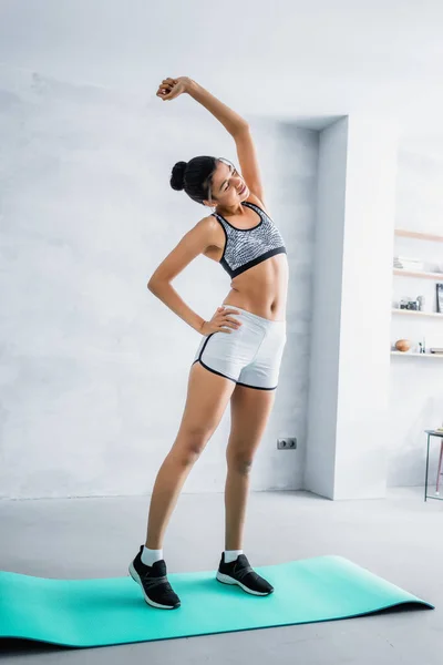 Vista completa della donna afro-americana in abbigliamento sportivo che fa esercizio di piega laterale sul tappeto fitness a casa — Foto stock