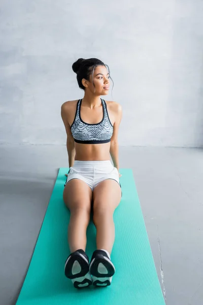 Junge afrikanisch-amerikanische Frau in Sportbekleidung schaut weg, während sie zu Hause auf einer Fitnessmatte sitzt — Stockfoto