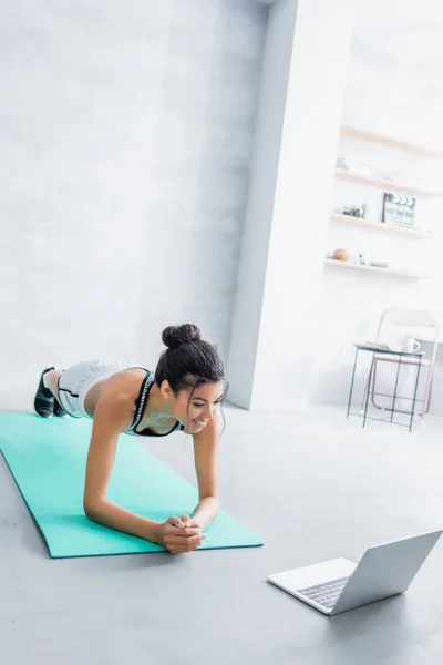 Joven afroamericana deportista haciendo ejercicio en la pose tablón en la alfombra de fitness cerca de la computadora portátil - foto de stock