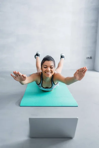 Cheerful african american sportswoman in locust pose near laptop on blurred foreground — Stock Photo