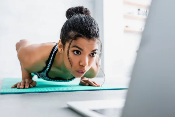 Afroamericana deportista haciendo flexiones ejercicio cerca de portátil en primer plano borrosa - foto de stock