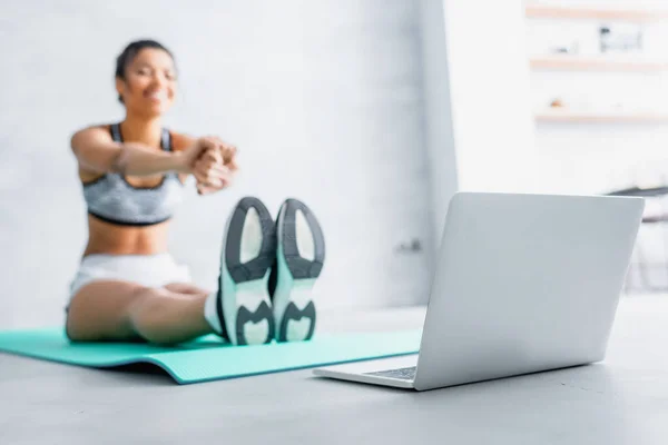 Jovem afro-americana esportista esticando os braços enquanto sentado no tapete de fitness perto do laptop, fundo borrado — Fotografia de Stock