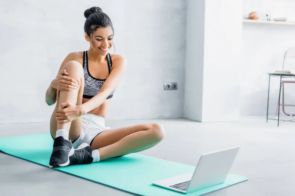 Young african american woman in sportswear smiling while sitting on fitness mat near laptop — Stock Photo