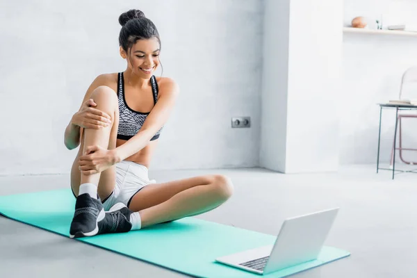 Junge Afroamerikanerin in Sportbekleidung lächelt, während sie in der Nähe des Laptops auf einer Fitnessmatte sitzt — Stockfoto