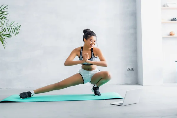 Улыбающаяся африканская американка делает упражнения на тренажерном коврике рядом с ноутбуком — стоковое фото
