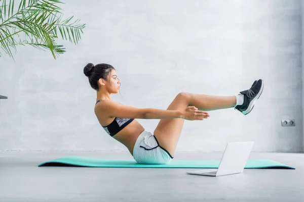Side view of african american sportswoman practicing half boat pose near laptop at home — Stock Photo