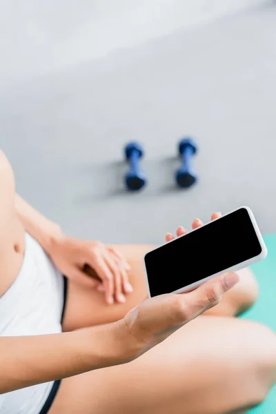 Cropped view of african american sportswoman holding smartphone with blank screen near dumbbells on blurred background — Stock Photo