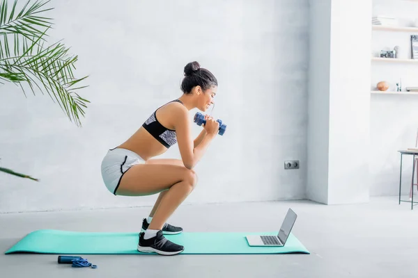 Side view of african american sportswoman doing sit ups with dumbbells near laptop at home — Stock Photo