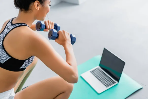 Cropped view of young african american sportswoman exercising with dumbbells near blurred laptop with blank screen — Stock Photo
