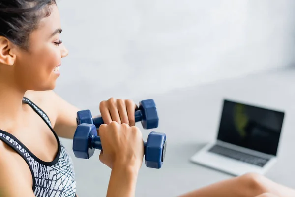 Young african american sportswoman exercising with dumbbells near laptop with blank screen on blurred background — Stock Photo