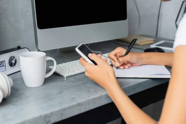 Vista recortada del freelancer afroamericano que sostiene el teléfono inteligente y escribe en un cuaderno - foto de stock