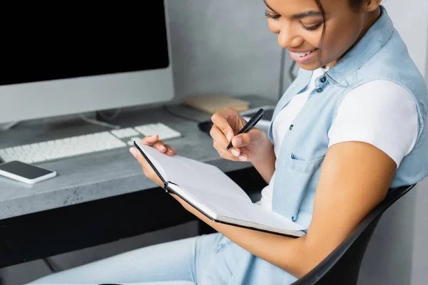 Sonriente freelancer afroamericano sosteniendo un cuaderno y una pluma cerca de un teléfono inteligente y una computadora sobre un fondo borroso - foto de stock