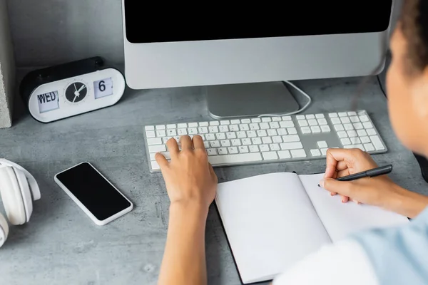 Visão recortada do freelancer afro-americano digitando no teclado e escrevendo no notebook perto do smartphone com tela em branco, foreground borrado — Fotografia de Stock