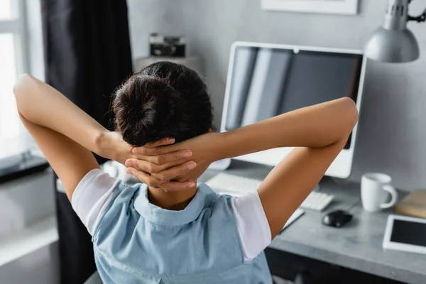 Back view of african american freelancer sitting with hands behind head near computer on blurred background — Stock Photo