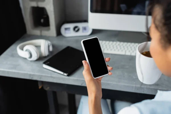 Vista ritagliata di afro-americano freelance con smartphone con schermo bianco vicino notebook e cuffie su sfondo sfocato — Foto stock