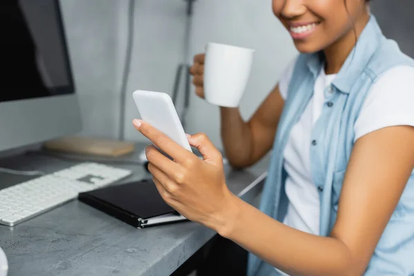 Vista recortada del freelancer afroamericano sosteniendo una taza de té y charlando en un teléfono inteligente, fondo borroso - foto de stock