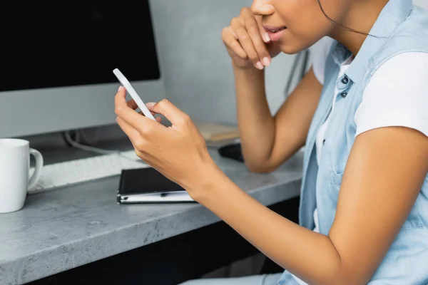 Ausgeschnittene Ansicht eines afrikanisch-amerikanischen Freelancers mit Smartphone in der Nähe des heimischen Schreibtisches — Stockfoto