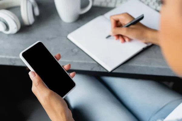 Partial view of african american woman holding smartphone with blank screen and writing in notebook, blurred foreground — Stock Photo