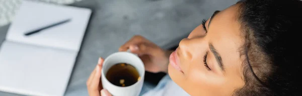 Vue aérienne de la femme afro-américaine tenant une tasse de thé chaud avec les yeux fermés, bannière, fond flou — Photo de stock