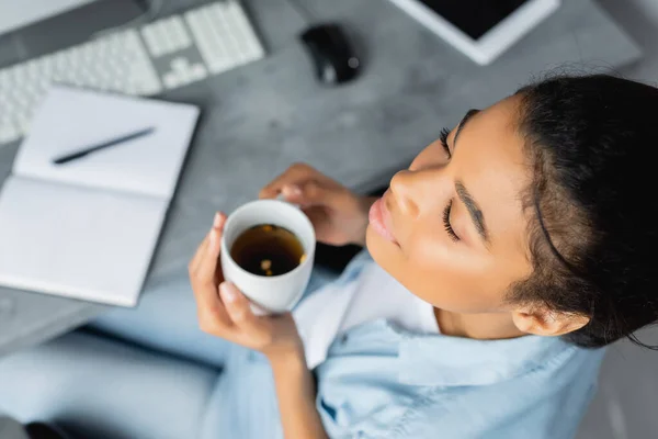 Overhead view of african american freelancer holding cup of tea while sitting with closed eyes, blurred background — Stock Photo