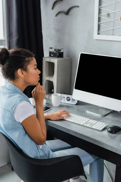 Joven afroamericano freelancer escribiendo en el teclado cerca del monitor de la computadora con pantalla en blanco - foto de stock