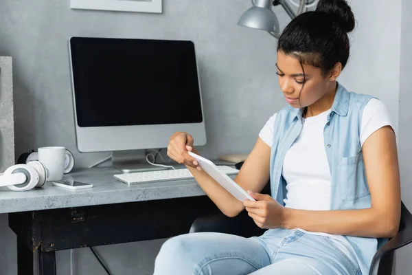 Joven afroamericano freelancer sosteniendo tableta digital mientras está sentado cerca de monitor con pantalla en blanco - foto de stock