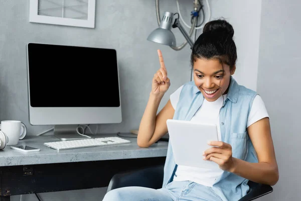 Emocionado freelancer afroamericano mostrando gesto de idea mientras sostiene la tableta digital cerca del monitor con pantalla en blanco - foto de stock