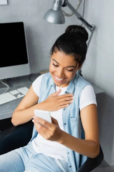 Cheerful african american freelancer touching chest while chatting on smartphone at home — Stock Photo