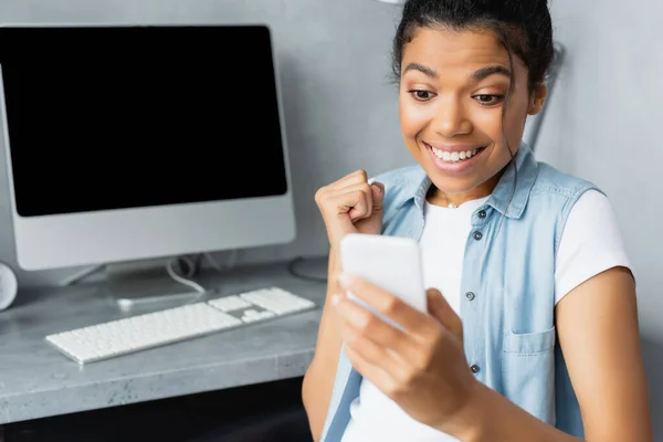 Emocionado freelancer afroamericano mostrando un gesto de victoria cerca del monitor de la computadora y el teclado en un fondo borroso - foto de stock