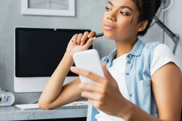 Riflessivo freelance afroamericano guardando altrove mentre tiene lo smartphone, in primo piano sfocato — Foto stock